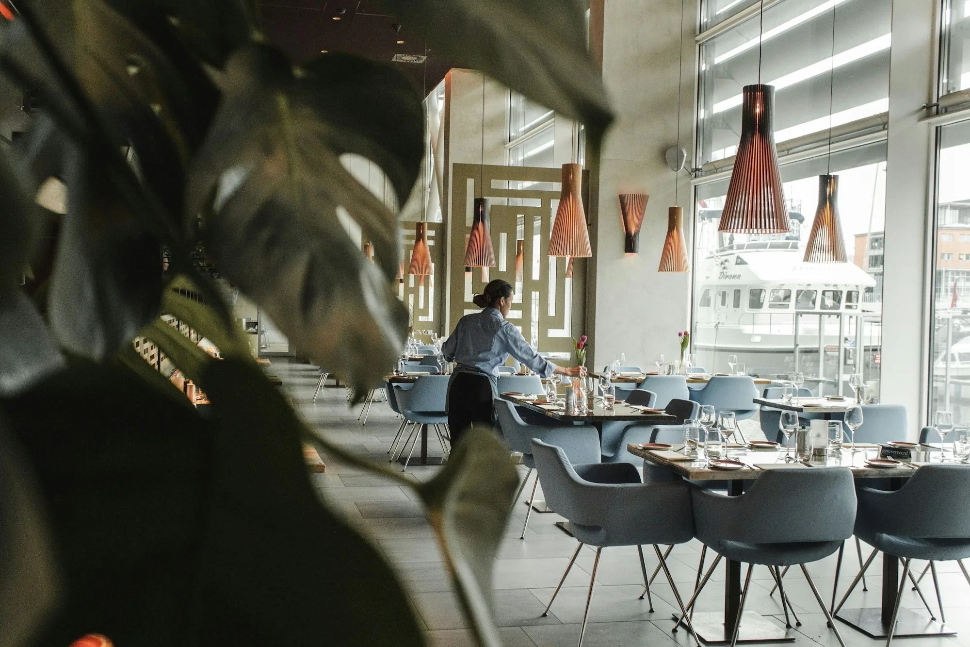 A restaurant manager setting tables in a hip waterfront dining room under copper pendant lights — the daily reality of running a restaurant floor